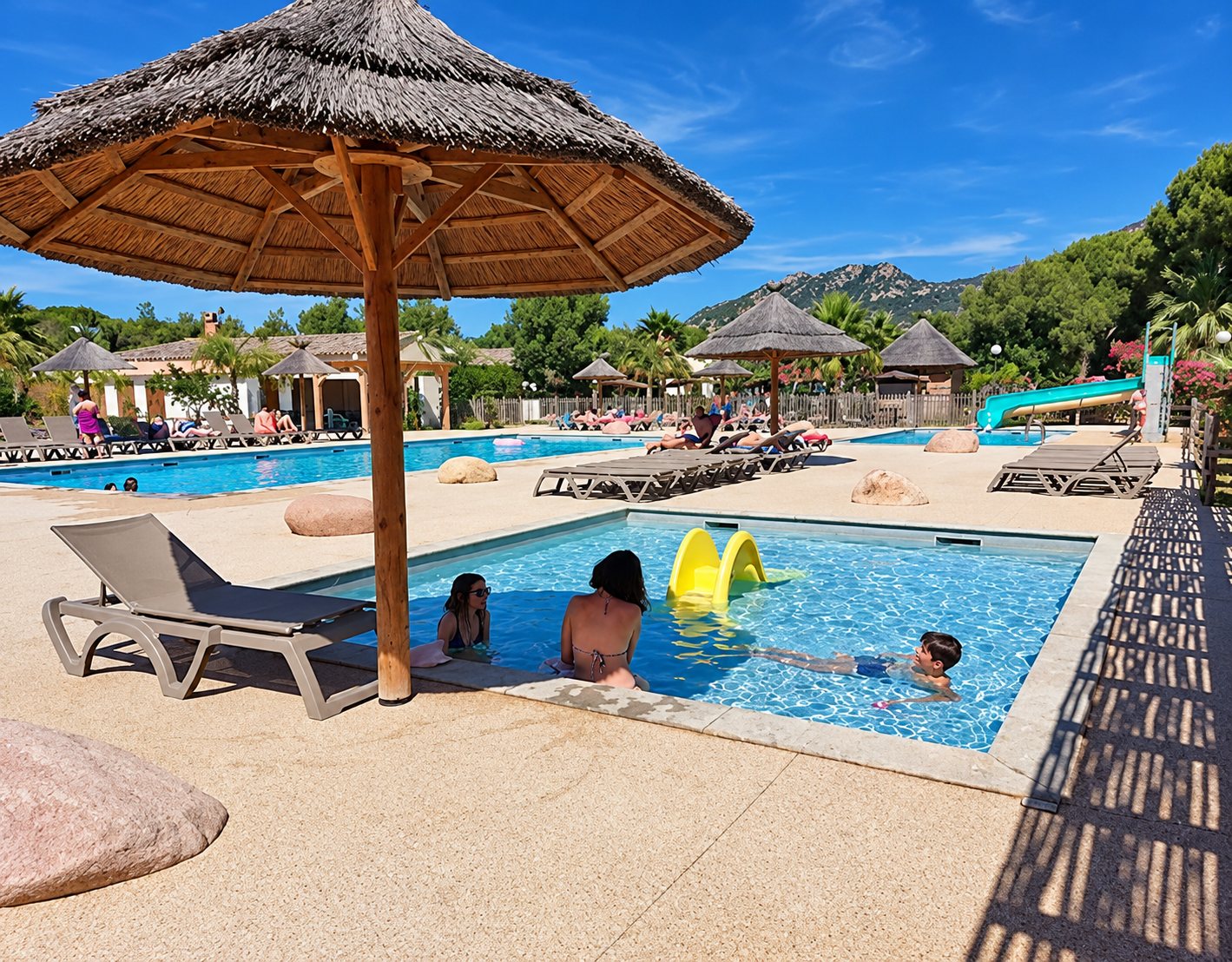 Des baigneurs profitent d'une piscine de camping entourée de transats et de parasols en paille, avec un toboggan et des montagnes en arrière-plan.
