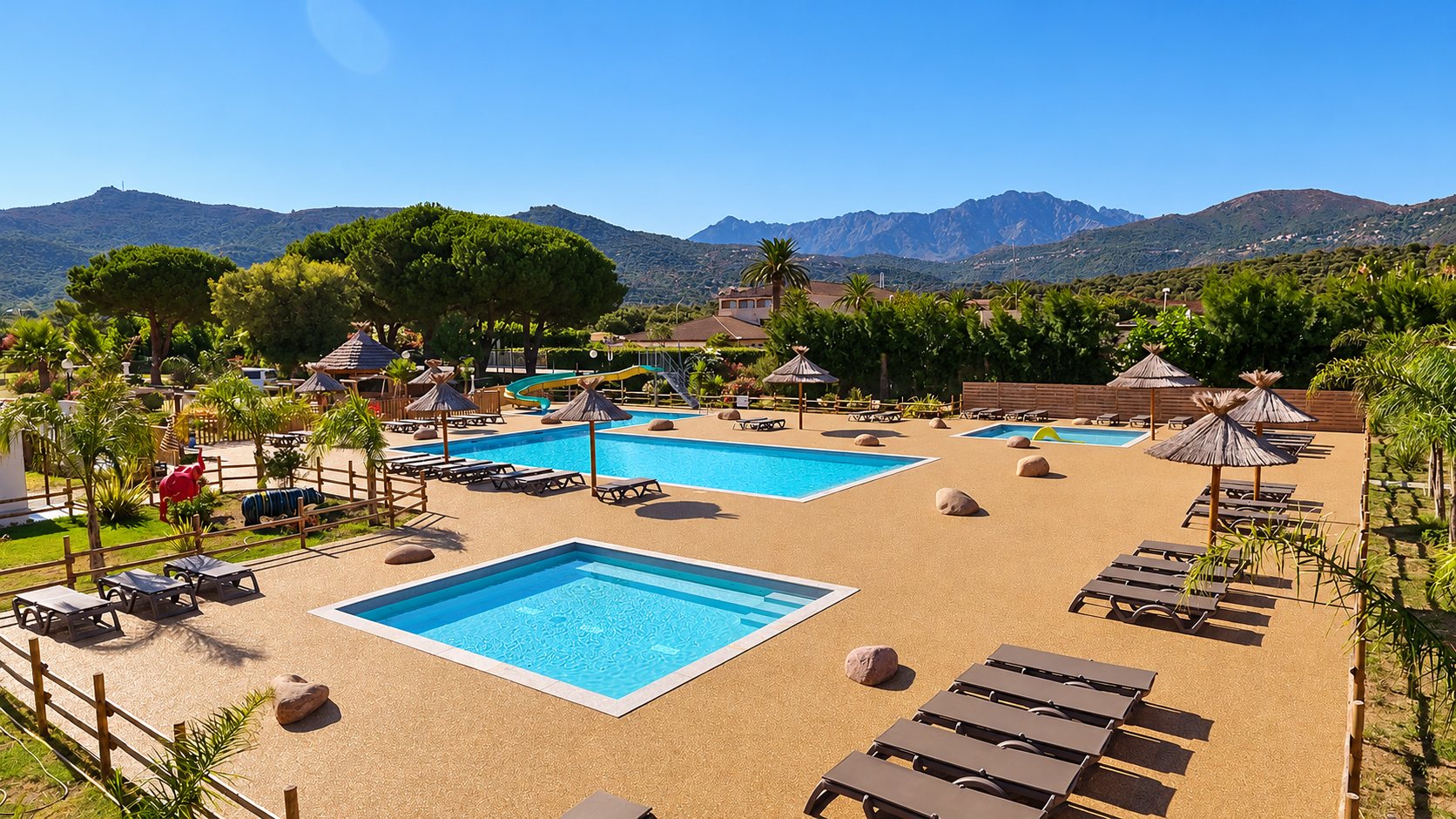 Vue aérienne d'un espace aquatique avec plusieurs bassins, des transats et des parasols en paille, entouré d'arbres et de montagnes sous un ciel dégagé.
