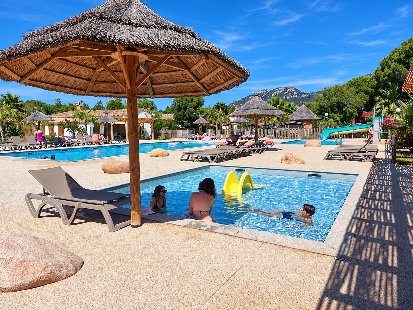 Des baigneurs profitent d'un bassin peu profond sous un parasol en paille, avec transats, grande piscine et toboggan en arrière-plan sous un ciel bleu.