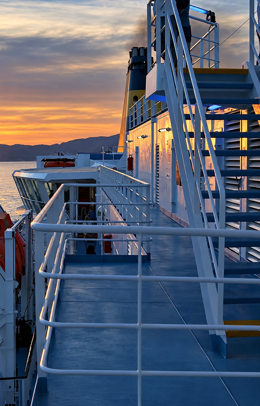 Le pont extérieur d'un ferry avec ses rambardes blanches et un escalier métallique, éclairé par la lumière du coucher de soleil au-dessus de la mer.