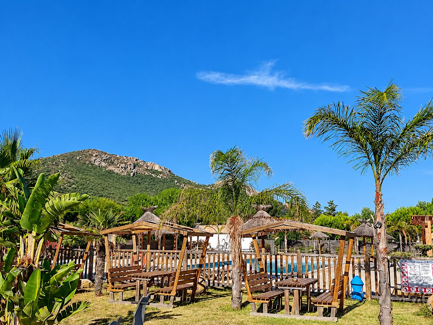 Des tonnelles en bois avec tables et bancs entourent une piscine bordée d'une clôture, au milieu de palmiers, avec une montagne et un ciel bleu en arrière-plan.